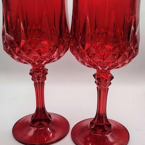 Könnte beinhalten: Zwei passende rote Weingläser aus Glas mit einem facettierten Rautenmuster im unteren Bereich des Kelches und einem strukturierten Stiel. Die Gläser haben einen breiten, runden Fuß und einen glatten, ausgestellten Rand in tiefem, durchscheinendem Rot.