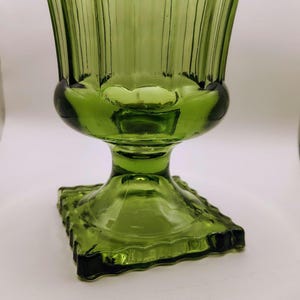May include: A green glass vase with a square base and a flared, ribbed top. The vase has a pedestal base and a bulbous midsection. The glass has a slight green tint and a glossy finish. The vase is likely vintage.