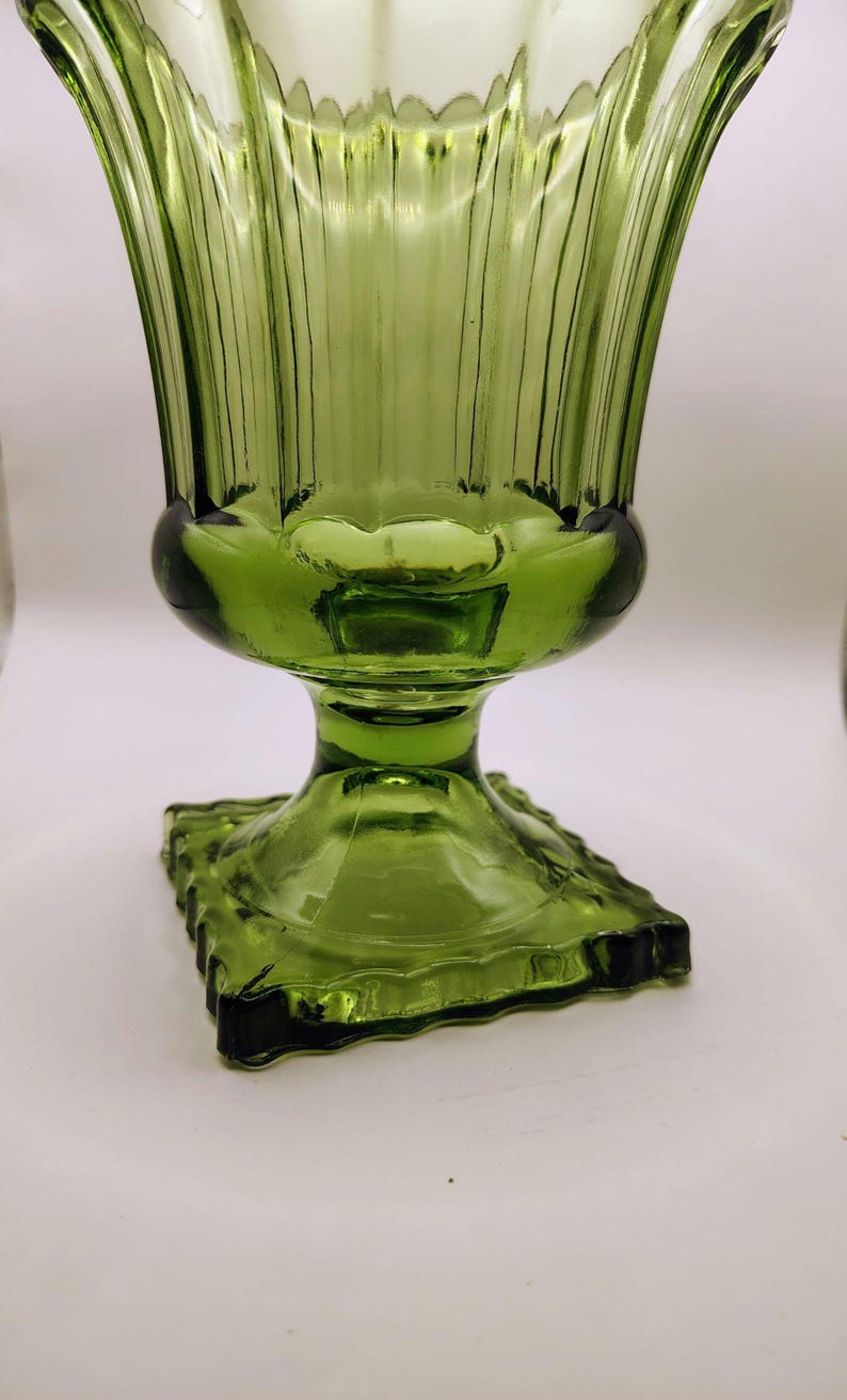 May include: A green glass vase with a square base and a flared, ribbed top. The vase has a pedestal base and a bulbous midsection. The glass has a slight green tint and a glossy finish. The vase is likely vintage.