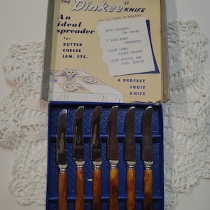 May include: A set of six stainless steel knives with amber-colored marbled handles, presented in a blue box. The box lid features text including 'The Dinkee Knife', 'An ideal spreader for butter, cheese, jam, etc.', and 'A perfect fruit knife'.