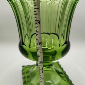 May include: Green glass vase with a scalloped rim and a square base. The vase is approximately 5 inches tall. The vase has vertical ridges and a flared shape. A measuring tape is placed next to the vase.