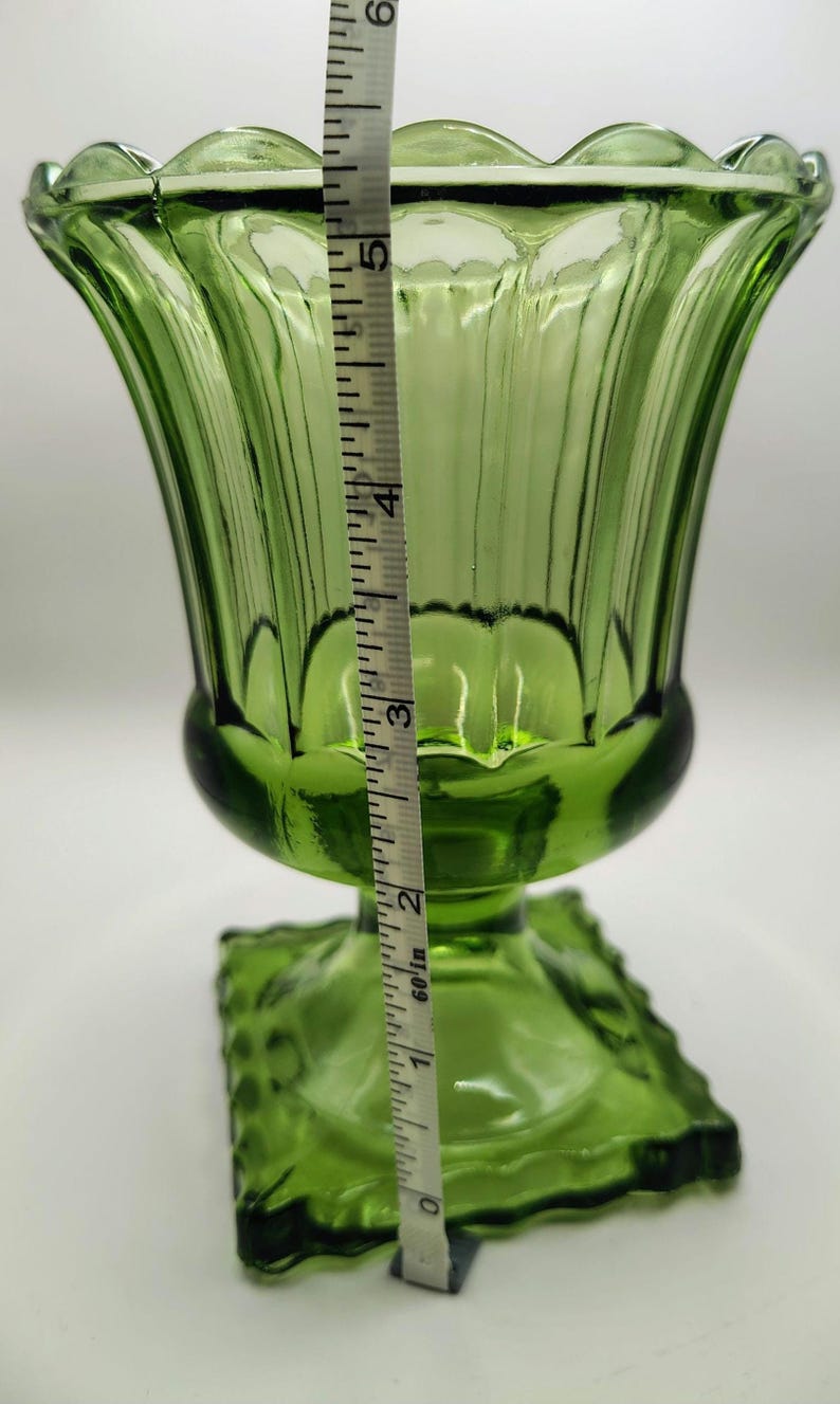 May include: Green glass vase with a scalloped rim and a square base. The vase is approximately 5 inches tall. The vase has vertical ridges and a flared shape. A measuring tape is placed next to the vase.
