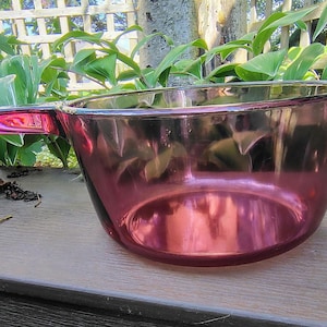 May include: A vintage, ruby-coloured glass saucepan with a handle. The saucepan is made of transparent glass, allowing the contents to be seen. The handle is a matching ruby colour. The saucepan is sitting on a wooden surface, with green foliage in the background.