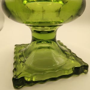 May include: A green glass pedestal bowl with a square base and a rounded top. The bowl has a textured, ribbed design and a stem. The glass is a vibrant, transparent green color.