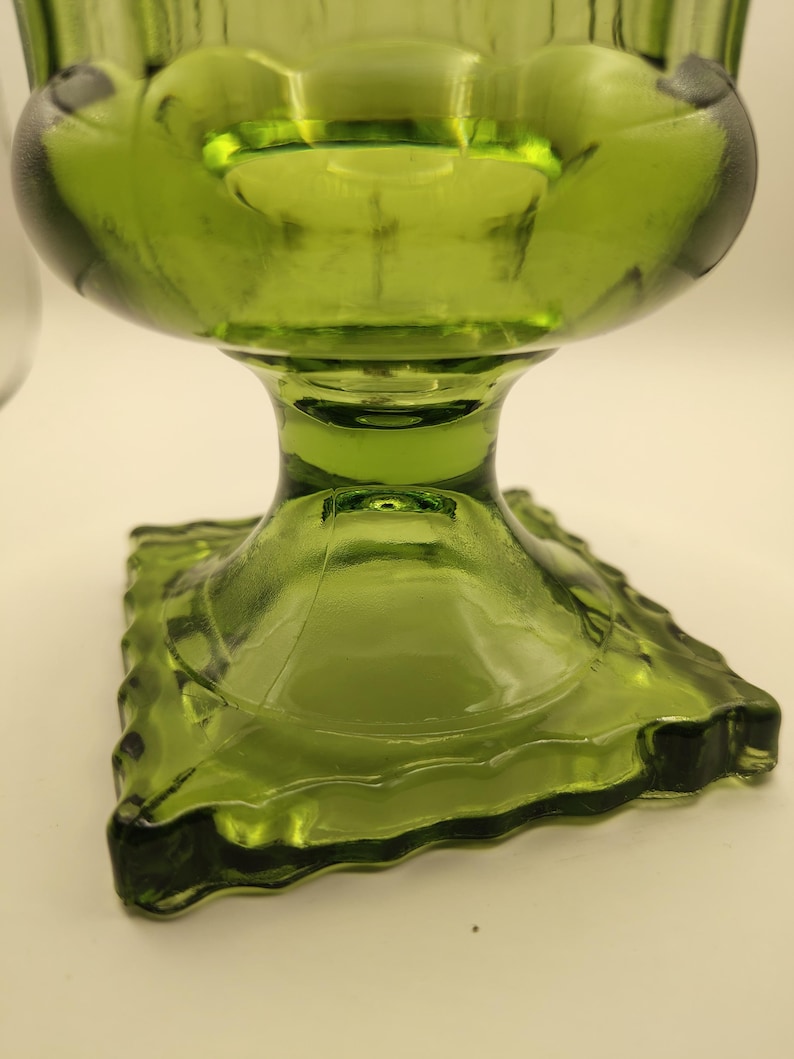 May include: A green glass pedestal bowl with a square base and a rounded top. The bowl has a textured, ribbed design and a stem. The glass is a vibrant, transparent green color.