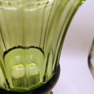 May include: Close-up of a green glass vase with a flared, ribbed design. The vase has a scalloped rim and a rounded base. The glass is transparent, allowing light to pass through, and has a glossy finish.