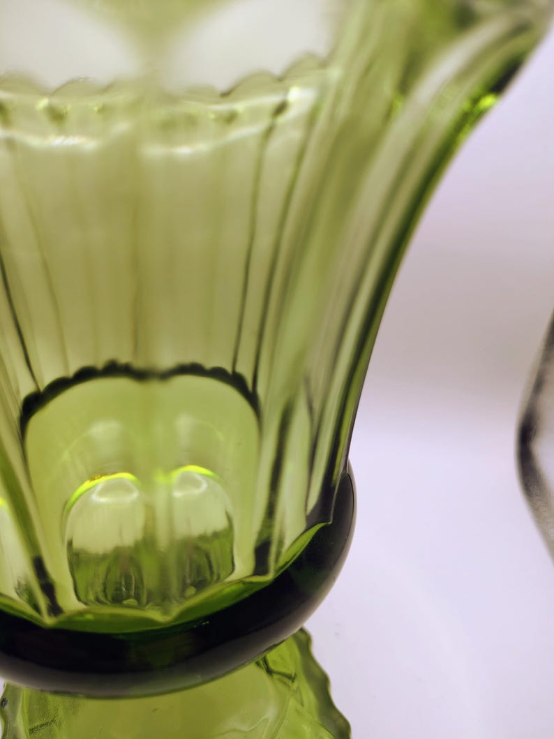 May include: Close-up of a green glass vase with a flared, ribbed design. The vase has a scalloped rim and a rounded base. The glass is transparent, allowing light to pass through, and has a glossy finish.