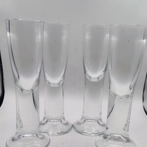 May include: Four clear, stemmed shot glasses. Each glass has a rounded base, a slender stem, and a tall, cylindrical bowl. The glasses are made of transparent glass, allowing light to pass through. They are arranged in a row against a white background.
