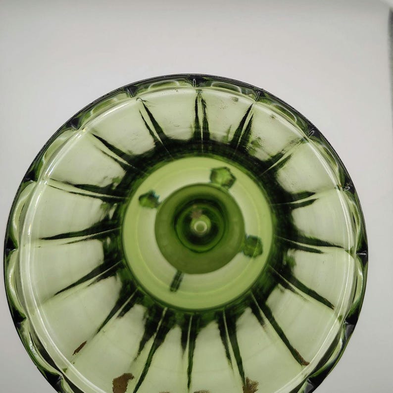 May include: An overhead view of a green glass object with a circular, ridged design. The object has a central stem and a series of radiating lines. The glass has a light green hue with darker green accents, creating a textured appearance.