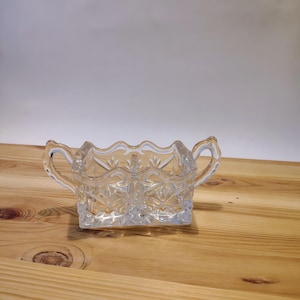 May include: A clear, cut-glass sugar bowl with two handles and a decorative, scalloped rim. The bowl features a diamond pattern and sits on a rectangular base. The sugar bowl is displayed on a light-coloured wooden surface.