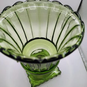 May include: A green glass vase with a scalloped rim and vertical ribbed detailing. The vase has a pedestal base with a geometric design. The glass is transparent, allowing light to pass through, and the overall shape is flared.
