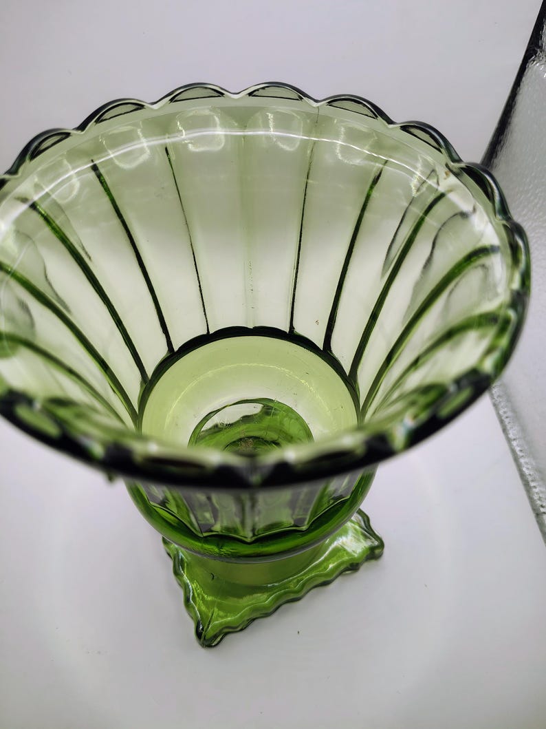May include: A green glass vase with a scalloped rim and vertical ribbed detailing. The vase has a pedestal base with a geometric design. The glass is transparent, allowing light to pass through, and the overall shape is flared.