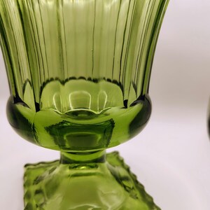 May include: A green glass vase with a fluted design. The vase has a square base and a rounded body that flares out at the top. The glass has a ribbed texture and a scalloped rim.