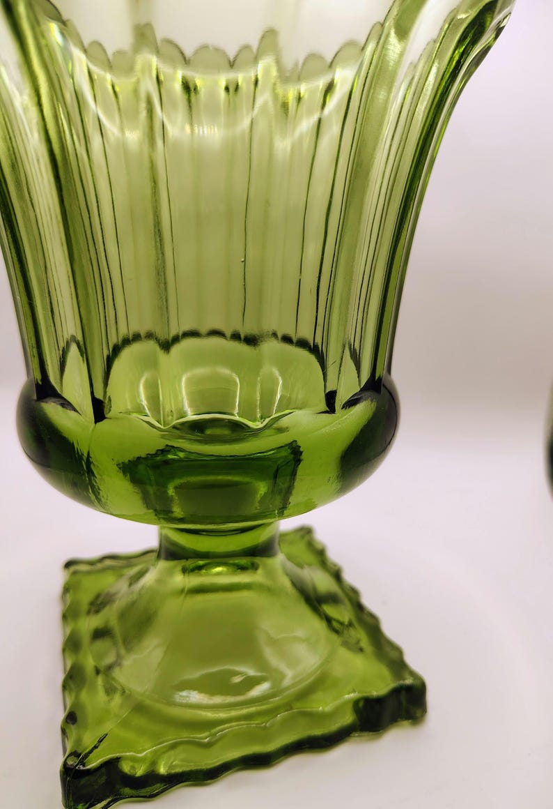 May include: A green glass vase with a fluted design. The vase has a square base and a rounded body that flares out at the top. The glass has a ribbed texture and a scalloped rim.