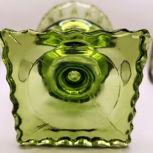 May include: A green glass pedestal dish with a square base and a ruffled edge. The dish has a circular design on the base and a textured surface. The glass is a vibrant shade of green.