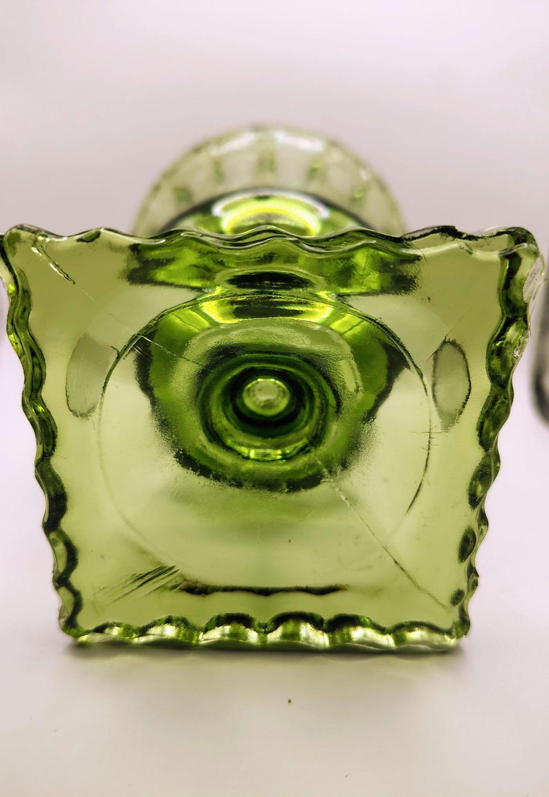 May include: A green glass pedestal dish with a square base and a ruffled edge. The dish has a circular design on the base and a textured surface. The glass is a vibrant shade of green.