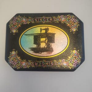 Könnte beinhalten: Schwarze rechteckige Dose mit ovaler Mitte, die eine Vintage-Illustration einer Singer-Nähmaschine zeigt. Das Wort "SINGER" ist in goldener Schrift über und unter dem Oval dargestellt. Florale und Scroll-Designs in Rosa, Gelb und Grün schmücken die Ecken.