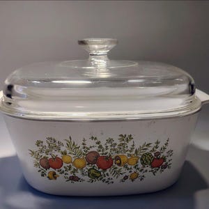 May include: White casserole dish with a clear glass lid. The dish features a colorful botanical design of tomatoes, lemons, and herbs. The lid has a clear glass knob. The dish is approximately 12 inches long.