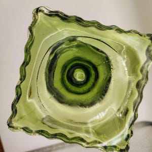 May include: A square, green glass bowl with a wavy, scalloped edge. The bowl's interior features concentric circles of varying shades of green, creating a textured, layered effect. The glass appears translucent.