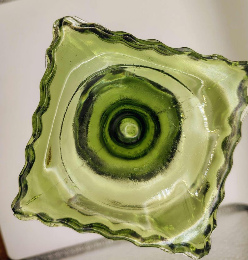 May include: A square, green glass bowl with a wavy, scalloped edge. The bowl's interior features concentric circles of varying shades of green, creating a textured, layered effect. The glass appears translucent.
