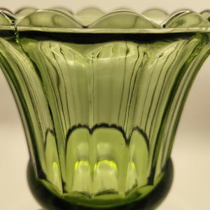 May include: A green glass vase with a scalloped rim and a flared, ribbed body. The vase has a rounded base and a square, textured foot. The glass has a slight green tint and a glossy finish.