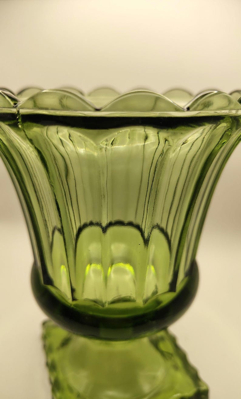 May include: A green glass vase with a scalloped rim and a flared, ribbed body. The vase has a rounded base and a square, textured foot. The glass has a slight green tint and a glossy finish.