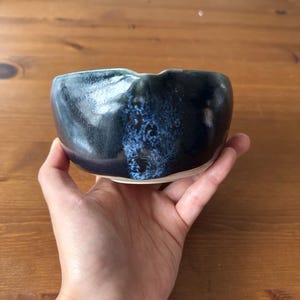 Könnte beinhalten: Eine handgefertigte Keramikschale mit dunkelblauer Glasur und hellblauem Innenraum. Die Schale hat einen unregelmäßigen Rand und einen weißen Boden. Die Schale wird in einer Hand gehalten, um ihre Größe und Form zu zeigen. Die Schale wird wahrscheinlich zum Servieren von Speisen oder als Dekorationsartikel verwendet.