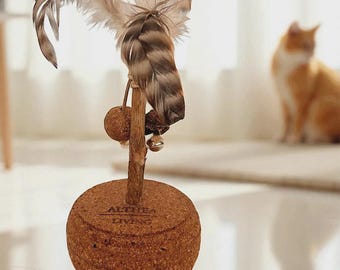 Puede incluir: Un juguete para gatos con una base de corcho y un palo de madera rematado con plumas. La base es una esfera marrón con el texto "ALTHEA LIVING". Las plumas son marrones y beige.