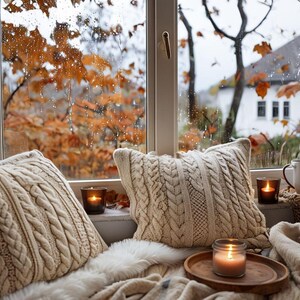 Puede incluir: Escena acogedora junto a la ventana con dos cojines de punto trenzado color crema, una manta y una bandeja de madera con una vela encendida. Una taza, una cesta y luces de cadena contribuyen al ambiente cálido y acogedor. La ventana da a las hojas de otoño.