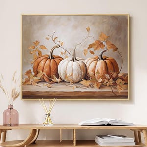 May include: A framed painting depicts three pumpkins in shades of orange and white, with autumn leaves. The pumpkins are arranged on a wooden surface, with a neutral background. The artwork is displayed above a wooden table with decorative items.