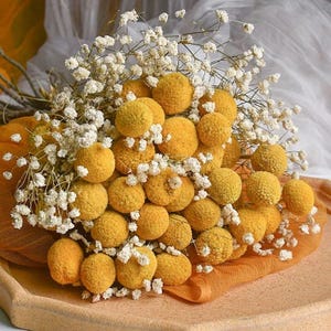 Könnte beinhalten: Ein Bouquet aus getrockneten gelben Billy Buttons und weißen Schleierkrautblüten. Das Arrangement befindet sich auf einem hellorangen Stoff und einem hellbraunen Tablett, was eine warme Ästhetik erzeugt.