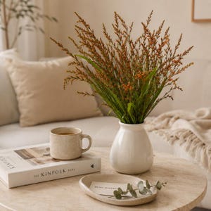 May include: A white ceramic vase filled with tall, slender stems of orange-tipped foliage. Beside the vase sits a speckled beige ceramic mug on a book titled The Kinfolk Home, and a small oval tray holding a card with the text Belle journée.