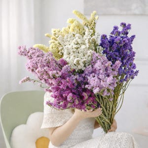 Peut inclure: Un grand bouquet de fleurs séchées aux tons violets, lavande, blancs et jaunes. Les fleurs sont tenues par une personne vêtue d'une robe en dentelle blanche. Le bouquet est un mélange de textures et de couleurs, créant un arrangement visuellement attrayant.