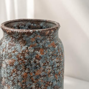 Könnte beinhalten: Eine zylindrische Keramikvase mit einer texturierten, gesprenkelten Oberfläche in einer Mischung aus tiefem Braun, erdigem Rost und hellblauem Glasur-Muster. Der Rand ist abgerundet und dick, mit einem rauen, porösen Finish am gesamten Körper.