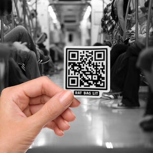 Puede incluir: Una pegatina blanca con un código QR negro y el texto "RAT BAG LIT" se sostiene frente a un vagón de metro. La pegatina es cuadrada con esquinas redondeadas. El fondo es una imagen en blanco y negro.