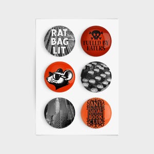 May include: A set of six round buttons with various designs. The buttons feature text like "RAT BAG LIT" and "ANTI SOCIAL BOOK CLUB", along with graphics of a rat, a skull, and a cityscape. The buttons are in shades of red, orange, black, and white.