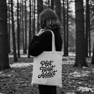 May include: A white tote bag featuring the text "Plot Twist Addict" in black script. The bag is held over the shoulder of a person wearing a black jacket, set against a backdrop of a forest.
