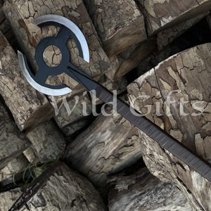 Könnte beinhalten: Eine schwarz-silberne Axt mit kreisförmigem Klingendesign und dunklem Griff. Die Axt liegt auf einem Haufen Holzscheite. Der Text "Wild Gifts" ist auf dem Bild zu sehen.