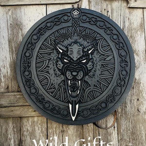 May include: A circular, black wall decoration featuring a carved wolf design. The wolf is depicted with an open mouth, surrounded by Celtic knotwork. The text "Wild Gifts" is at the bottom. It is mounted on a wooden door.