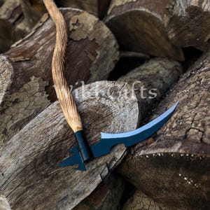 Puede incluir: Una guadaña artesanal con una hoja curva negra y un mango de madera. El mango tiene un diseño tallado y retorcido. La guadaña descansa sobre una pila de troncos de madera desgastados. El texto "Wild Gifts" es visible.