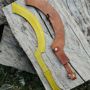 Könnte beinhalten: Ein gelbes Kukri-Messer mit gebogener Klinge und Griff, neben einer braunen Lederscheide. Das Messer liegt auf einer verwitterten Holzoberfläche, die Scheide ist mit einem Knopf und Riemen befestigt.