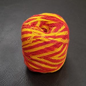 May include: A round ball of yarn featuring a vibrant, alternating pattern of bright yellow and deep red strands. The fibres are tightly wound, creating a textured, spherical shape with visible individual threads throughout the entire surface.