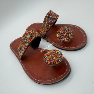 May include: A pair of brown leather sandals with colorful beaded accents. The sandals feature a strap across the top of the foot and a circular beaded design on the toe. The sandals are handmade and have white stitching.