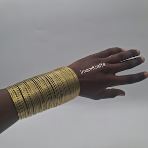 May include: A large stack of thin, circular gold-toned metal bangles worn on the wrist. The bangles have a smooth, reflective surface and a uniform, slender profile. The text ImaniKrafts is visible in white lettering near the wrist area.