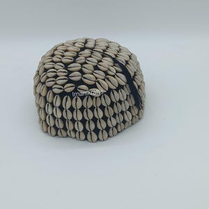 Peut inclure: Un bonnet beige et noir orné de petits coquillages blancs. Les coquillages sont disposés de manière serrée, créant une surface texturée. Le bonnet est de forme arrondie, avec une bande noire à la base. La marque "Imanikrafts" est visible.
