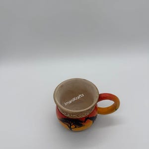 Puede incluir: Taza de cerámica pintada a mano con un vibrante diseño de puesta de sol. La taza presenta una silueta negra de un árbol sobre un fondo amarillo y naranja. El asa es naranja y el interior es de color marrón claro. El texto "ImaniKrafts" es visible.