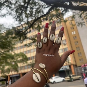 May include: A hand displays a collection of gold-toned jewelry featuring cowrie shells. The set includes multiple rings and a cuff bracelet. The nails are painted red. The brand name "imanikrafts" is visible.