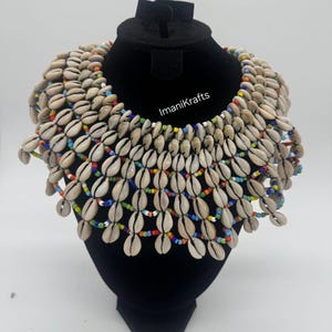 May include: A statement necklace made of numerous beige seashells, strung together with colorful beads. The necklace is displayed on a black mannequin head. The text "ImaniKrafts" is visible on the mannequin.