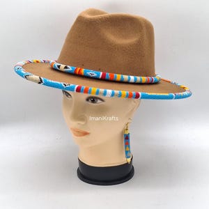 Puede incluir: Un sombrero fedora marrón con una banda de cuentas en azul, blanco, naranja y amarillo. El sombrero se exhibe en una cabeza de maniquí, con pendientes de cuentas a juego. El texto "ImaniKrafts" es visible.