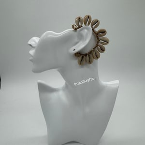 May include: A unique ear cuff featuring a cluster of small, beige seashells arranged in a circular design. The shells are attached to a gold-colored wire frame. The ear cuff is displayed on a white mannequin head.